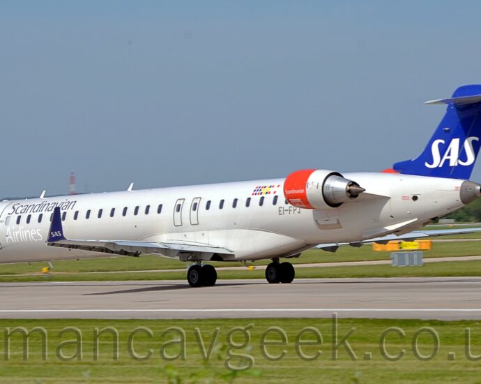Side view of a long, thin, white, twin engined jet airliner with the engines mounted on the sides of the rear fuselage. The plane is moving from right to left and slightly away from the camera on a grey runway. The plane is almost entirely white, with silver "Scandinavian" titles on the upper forward fuselage, just above the first few passenger cabin windows, with even whiter "Airlines" titles on the lower forward fuselage, under the windows. The registration "EI-FPJ" is on the lower rear fuselage, under the last of the windows, just in front of the red engine pods. The tail is a deep blue, with white "SAS" titles. The engine pods are red at the front, with small white "Scandinavian" titles. The upturned wingtips are the same blue as the tail, with the same "SAS" logo. Green grass fills the foreground, with the odd stray branch of small trees at the bottom. More grass leads off to trees lining the airfield perimeter in the distance, under grey-blue skies.