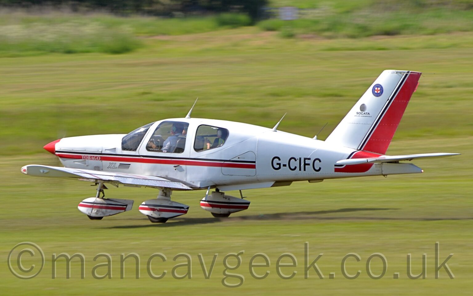 Side view of a single engined light aircraft moving at speed from right to left along a grass runway, with the nose wheel lifted slightly off the ground, and a small amount of flaps hanging from the rear of the wings, suggesting it is taking off. The plane is white, with a thin black and thicker red stripe running along the body, overlaid with the black registration "G-CIFC" on the rear fuselage. The spats on the undercarriage are white, with a similar black and red stripe. Green grass fills the rest of the frame.