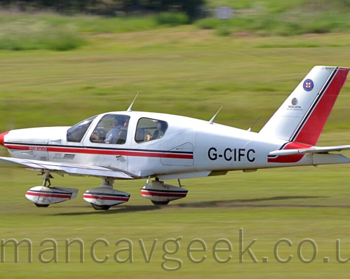 Side view of a single engined light aircraft moving at speed from right to left along a grass runway, with the nose wheel lifted slightly off the ground, and a small amount of flaps hanging from the rear of the wings, suggesting it is taking off. The plane is white, with a thin black and thicker red stripe running along the body, overlaid with the black registration "G-CIFC" on the rear fuselage. The spats on the undercarriage are white, with a similar black and red stripe. Green grass fills the rest of the frame.