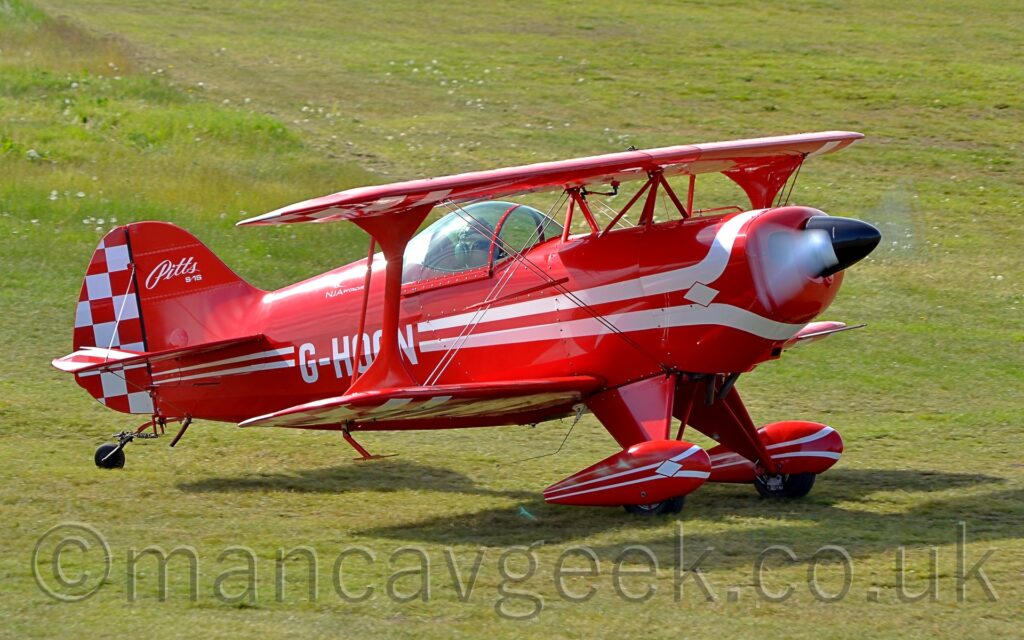 Side view of a red, single-engined biplane taxiing from left to right on a grass airfield. A pair of white lines wrap above and below the engine cowling in the nose, then run towards the rear, getting thinner as they go, overlaid with the white registration "G-HOON" on the lower rear fuselage, just aft of the bubble canopy over the single seat cockpit. The text "Pitts S-1S" is on the top half of the front of the red tail, with a red and white checkerboard pattern on the rear. The checkerboard pattern is repeated on the outer faces of the undersides of the wings. The white propellor at the front is a spinning white blur. Red spats over the main wheels have similar white stripes on the side Green grass fills the rest of the frame.