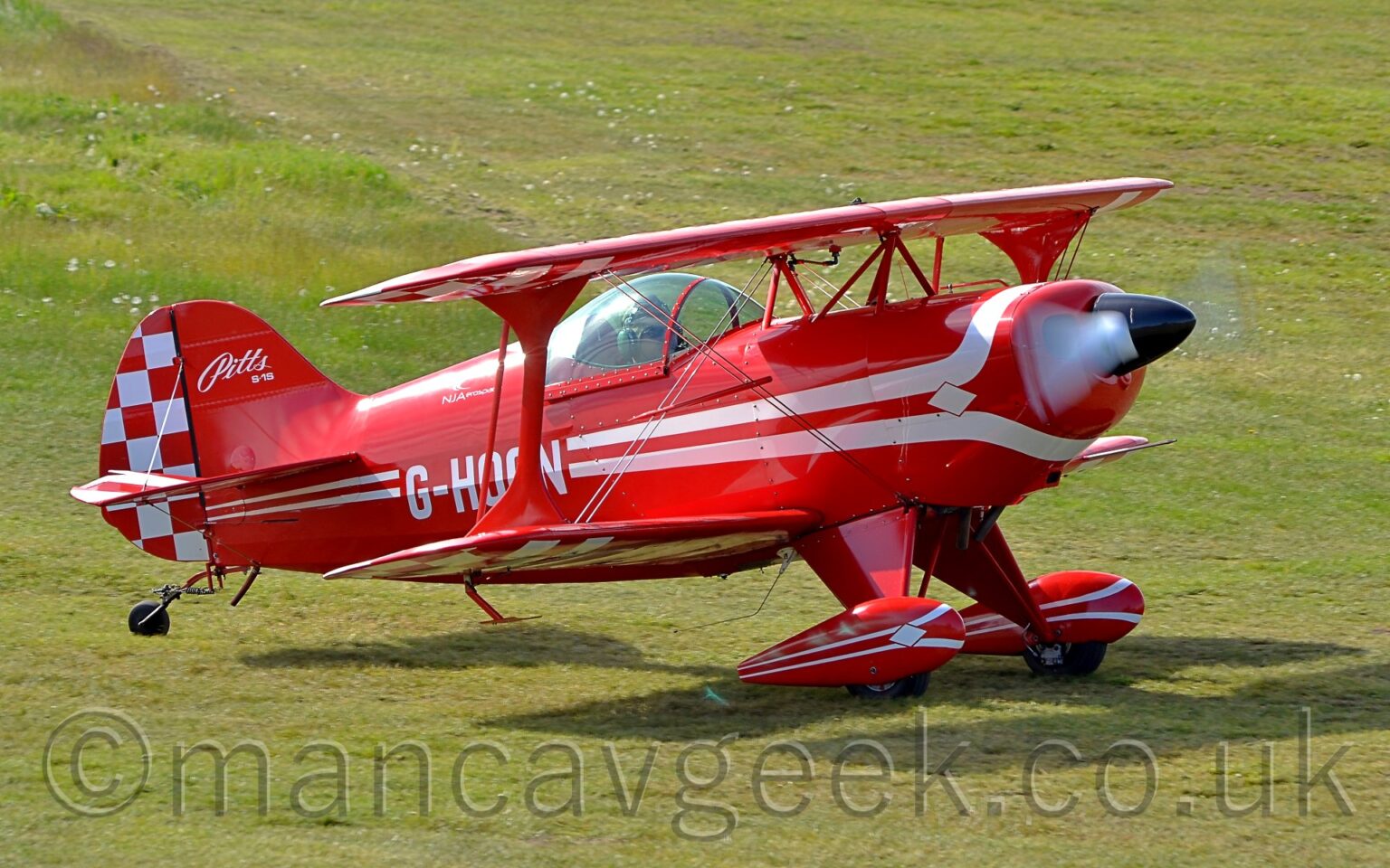 Side view of a red, single-engined biplane taxiing from left to right on a grass airfield. A pair of white lines wrap above and below the engine cowling in the nose, then run towards the rear, getting thinner as they go, overlaid with the white registration "G-HOON" on the lower rear fuselage, just aft of the bubble canopy over the single seat cockpit. The text "Pitts S-1S" is on the top half of the front of the red tail, with a red and white checkerboard pattern on the rear. The checkerboard pattern is repeated on the outer faces of the undersides of the wings. The white propellor at the front is a spinning white blur. Red spats over the main wheels have similar white stripes on the side Green grass fills the rest of the frame.