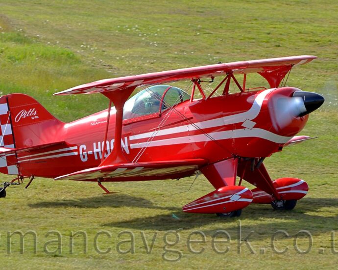 Side view of a red, single-engined biplane taxiing from left to right on a grass airfield. A pair of white lines wrap above and below the engine cowling in the nose, then run towards the rear, getting thinner as they go, overlaid with the white registration "G-HOON" on the lower rear fuselage, just aft of the bubble canopy over the single seat cockpit. The text "Pitts S-1S" is on the top half of the front of the red tail, with a red and white checkerboard pattern on the rear. The checkerboard pattern is repeated on the outer faces of the undersides of the wings. The white propellor at the front is a spinning white blur. Red spats over the main wheels have similar white stripes on the side Green grass fills the rest of the frame.