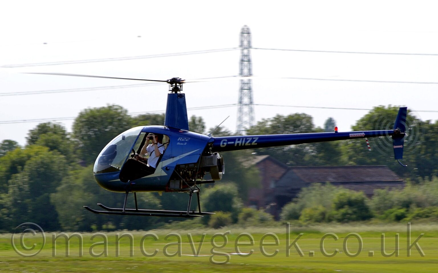 Side view of a small helicopter hovering a few metres above a grassy field, facing to the left. The copter is in a metallic blue livery, with a large golden zig-zag stripe on the side of the fuselage, partially missing because the doors aren't being carried on this flight, and the golden registration "G-GHIZZ" on the long, thin tail boom. A man is sitting in the left seat in the cockpit, holding a camera as he takes a photo towards the viewer. Green grass in the background leads up to trees stretching across the frame in the distance, and a tall electricity pylon with cables that reach across the frame, under cloudy white sky.