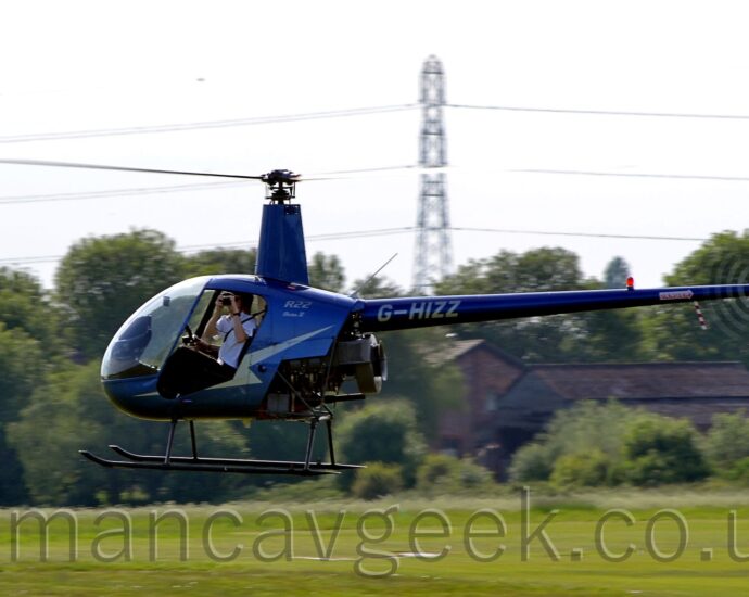 Side view of a small helicopter hovering a few metres above a grassy field, facing to the left. The copter is in a metallic blue livery, with a large golden zig-zag stripe on the side of the fuselage, partially missing because the doors aren't being carried on this flight, and the golden registration "G-GHIZZ" on the long, thin tail boom. A man is sitting in the left seat in the cockpit, holding a camera as he takes a photo towards the viewer. Green grass in the background leads up to trees stretching across the frame in the distance, and a tall electricity pylon with cables that reach across the frame, under cloudy white sky.