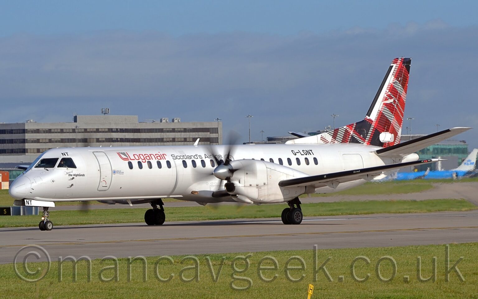 Side view of a white, twin propellor-engined airliner parked on a runway facing to the left, it's propellors blurred as they spin. The plane has red "LoganAir" titles on the upper forward fuselage, followed by black text "Scotlands Airline". The registration "G-LGNT" is on the upper rear fuselage, in black. The tail is covered in a red, black, and white tartan pattern. Green grass fills the foreground, with more grass in the background leading up to a large grey building in the distance. Pale blue sky with smears of grey cloud fill the rest of the frame