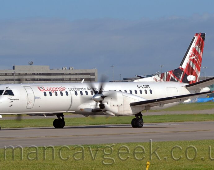 Side view of a white, twin propellor-engined airliner parked on a runway facing to the left, it's propellors blurred as they spin. The plane has red "LoganAir" titles on the upper forward fuselage, followed by black text "Scotlands Airline". The registration "G-LGNT" is on the upper rear fuselage, in black. The tail is covered in a red, black, and white tartan pattern. Green grass fills the foreground, with more grass in the background leading up to a large grey building in the distance. Pale blue sky with smears of grey cloud fill the rest of the frame