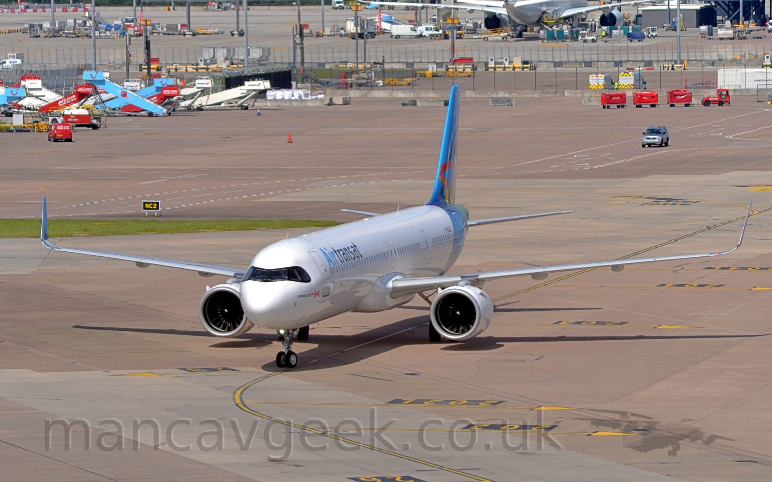 Side view of a twin engined jet airliner taxiing from right to left. The plane is mostly white, with a blue rear fuselage and tail, and 2 tone blue "Air Transat" titles on the upper forward fuselage. Grey concrete apron fills most of the background, with various pieces of airport furniture -mobile air stairs, lighting poles, luggage carts, and more - littering the area, and another airliner in the top right corner, with trees in the distance.
