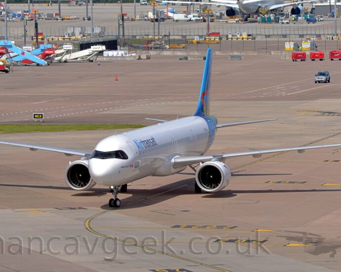 Side view of a twin engined jet airliner taxiing from right to left. The plane is mostly white, with a blue rear fuselage and tail, and 2 tone blue "Air Transat" titles on the upper forward fuselage. Grey concrete apron fills most of the background, with various pieces of airport furniture -mobile air stairs, lighting poles, luggage carts, and more - littering the area, and another airliner in the top right corner, with trees in the distance.