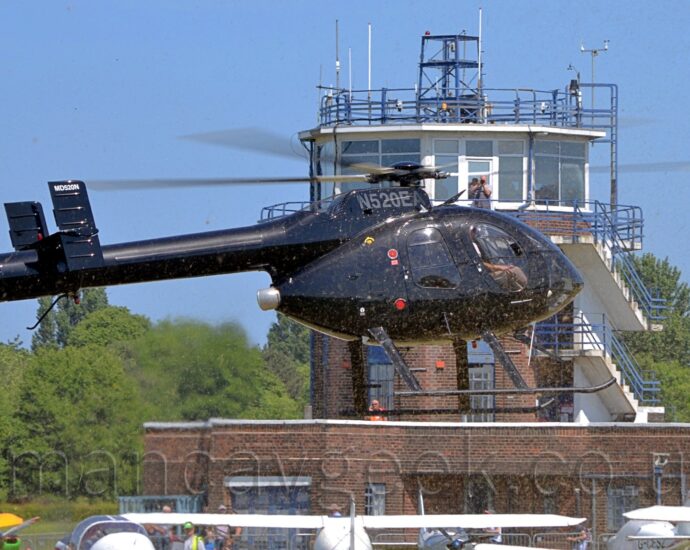 A black helicopter hovering several metres above the ground. There are very few markings, the silver registration "N520EA" on the engine pod on top of the fuselage, and the white text "MD520N" at the top of the twin tails at the end of the long tail boom. Mown grass flies in the air being forced down by the large rotor blades. Several white light aircraft can be seen in the background at the bottom of the frame, a background dominated by a brown brick control tower with a heavily glazed white top floor, where a man standing on a balcony is taking of photo of the helicopter. Trees stretch across the frame in the distance, under blue skies.