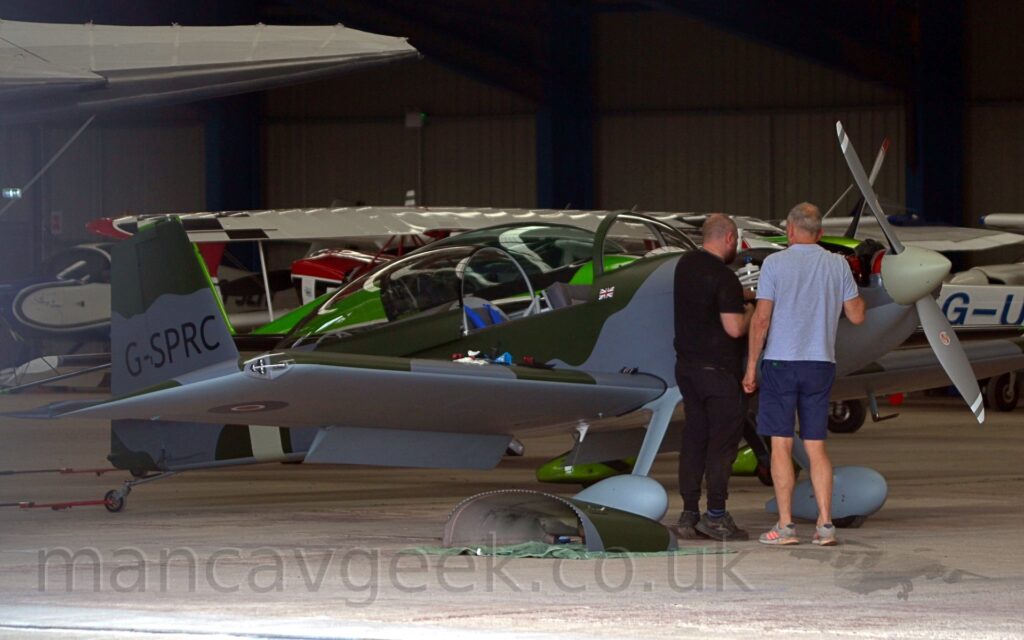 Side view of a single engined light aircraft parked facing to the right in a grey hangar. The plane is covered in a grey and olive green camouflage scheme on the fuselage and upper surfaces of the wings, with the belly and undersides of the wings painted a flat grey. A blue, white, and red roundel, outlined in yellow, can be seen under the planes right wing, with the black registration "G-SPRC" on the tail. The large bubble canopy over the cockpit is slid backwards revealing the blue seat-belt straps. A couple of men are leaning over the open engine cowling, looking at the engine bay. More planes can be seen in the background, further in the hangar.