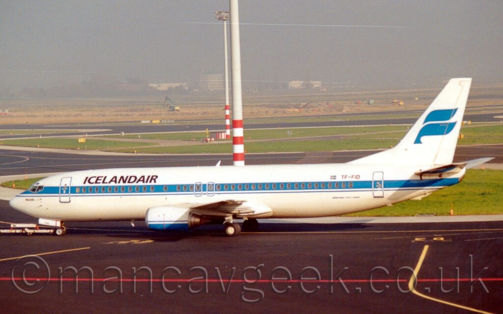 Side view of a twin engined jet airliner facing to the left but being pushed back to the right by an out-of-frame tug attached to the nose wheel by a long white pole. The plane is mostly white, with a blue stripe running along the body, covering the passenger cabin windows, and black "IcelandAir" titles on the upper forward fuselage. The black registration is on the upper rear, next to a small Icelandic flag, with black text "Boeing 737-400" on the lower rear. The white tail has a pair of blue twisted ribbons, vaguely resembling a letter "F". Black taxiway marked with red and yellow lines fill the foreground, with more black taxiways wending their way through islands of green grass, outlined by grey concrete, some of which have tall lighting poles with red and white banded bases reaching out of the top of the frame, in the background. Trees and tall buildings are slowly being swallowed up by grey haze in the distance.