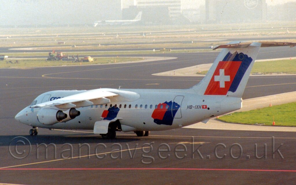 Side view of a high-winged, 4 engined jet airliner taxiing from right to left along a black taxiway marked with red and yellow lines. The plane is mostly white, with a small red splodge slightly offset to the right of a larger dark blue splodge under the wings, on the rear fuselage, and on the tail, the latter of which has the red splodge replaced with a red quadrilateral with a white cross in the middle. The black registration "HB-IXH" is on the upper rear fuselage, next to a small white Swiss flag. Black taxiway fills the foreground, with large green grass islands surrounded by more black taxiways in the background, with buildings and another plane in the distance being swallowed up in grey haze.