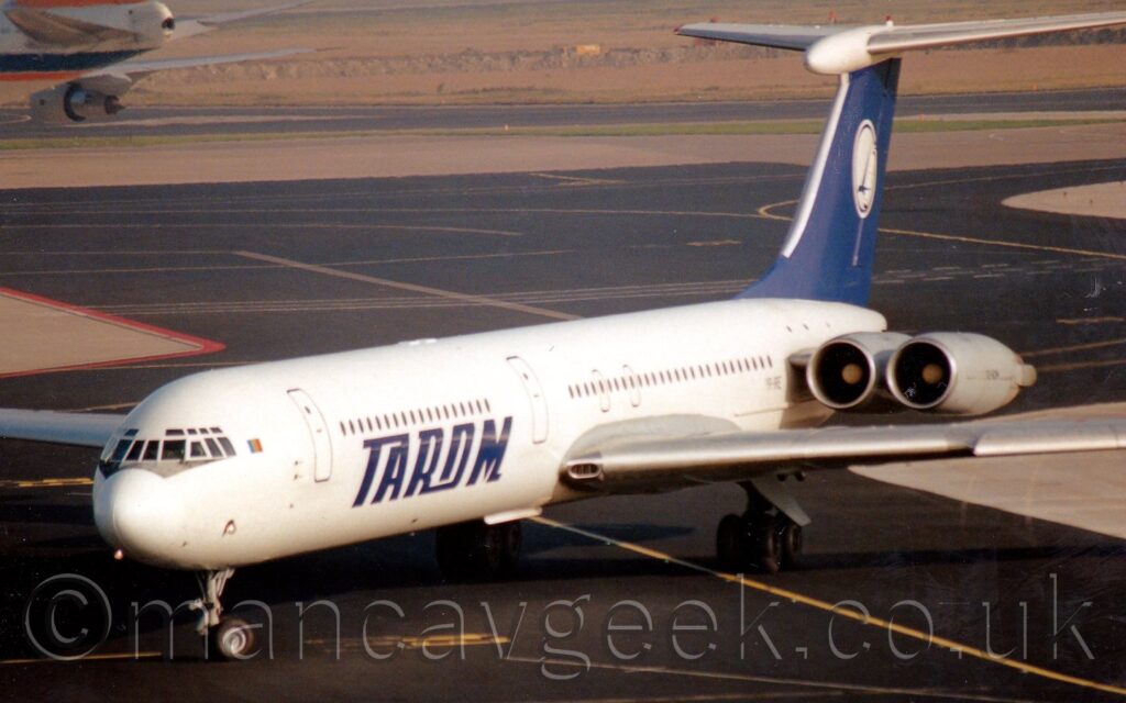 Side view of a white, 4 engined jet airliner with the engines mounted in pairs on the sides of the rear fuselage. There are large white "Tarom" titles on the sides of the lower fuselage. The tail is blue, with a large white circle in the middle containing a blue flying bird. Black tarmac taxiways fill the foreground and most of the background, with wide strips of grey concrete marking the edges of the taxiways, often with grassed areas beyond the concrete. The rear fuselage of a very large jet airliner with an orange tail can be seen in the top left corner, taxiing out of frame.