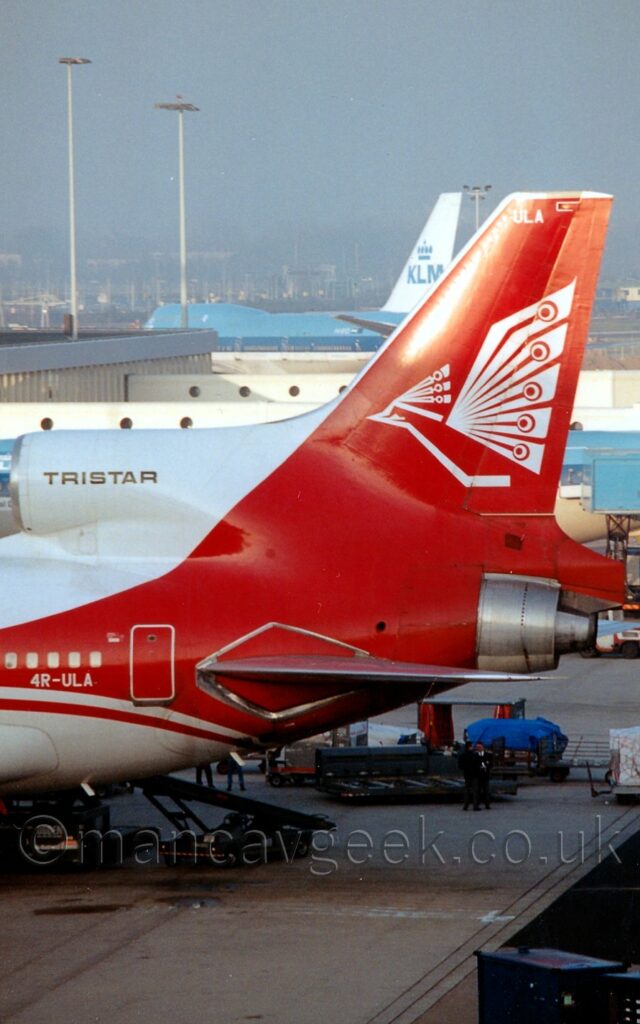 Close-up of the tail and rear fuselage of a 3 engined jet airliner parked facing to the left, nose-in to a terminal building. The plane is largely white, with a red stripe running along the body, covering the passenger cabin windows, before reaching up to fill the tail and down to fill the rear fuselage. A stylised, white bird with outstretched wings fills the red tail, with the white letters "ULA" right at the top, matching the white registration "4R-ULA" on the lower rear fuselage. Grey concrete apron fills the foreground, with blue cargo loaders with some baggage containers are visible under the planes body. A terminal building stretches into frame from the left, with white airbridges reaching out to a couple of blue and white planes in the background. Hazy grey sky fills the rest of the frame.