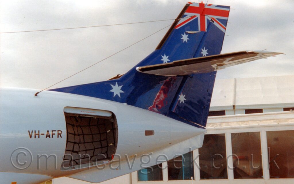 Side view of the rear fuselage and tail of a twin propellor-engined airliner parked facing to the right with the cargo hold door open. The plane is mostly white, with a dark blue tail with 5 white stars scattered around, and part of a British flag at the top. The black registration "VH-AFR" is in front of the open cargo door. In the background, a heavily glazed, low white building is on the lower right of the frame, with flat white sky fills the rest of the frame.
