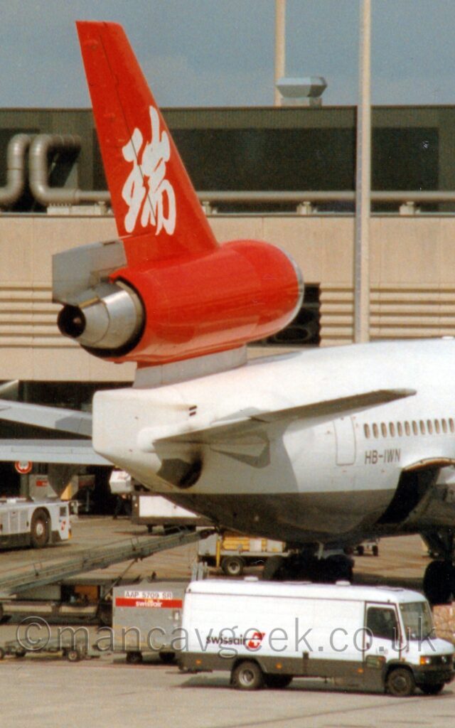 Close-up of the rear fuselage and tail of a 3 engined jet airliner parked facing to the right and away from the camera, nose-in to a brown terminal building. The plane's body is white, with a silvery-grey belly, with the black registration "HB-IWN" on the lower rear fuselage, between an open rear cargo hold and the rear passenger door. The red tail has large, white, Chinese writing in the middle. In the foreground, a white truck with black "SwissAir" titles on the lower rear is parked in front of this plane, with a grey cargo container sitting behind it. A sandy brown terminal building stretches across the frame, under grey-blue sky.
