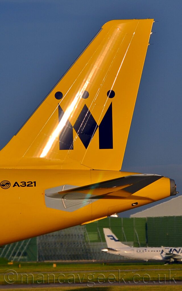 Close-up of the bright yellow rear fuselage and tail of a jet airliner facing to the left. There is black text "A321" next to a circular black logo on the rear fuselage, and a large, black letter "M" made to look like a crown on the tail. Green grass under the tail in the background leads up to a boxy, green metal frame, with a white jet airliner taxiing past in front. Blue sky fills the rest of the frame.