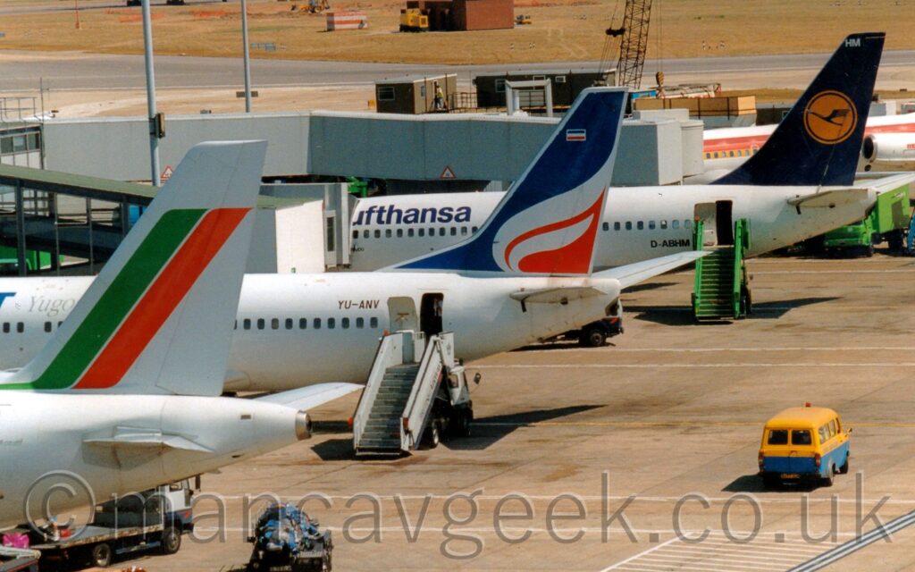 A line-up of medium-range airliners parked by a terminal building, all facing to the left, with their tails displayed prominently. Nearest the camera, on the left, is the white rear fuselage and tail of a jet airliner, with a diagonal green and red stripe on the tail. Next is a white jet airliner, with a blue and red tail with a white flame in the middle, and a set of white airstairs by the open rear door. At the rear is a white jet airliner with blue "Lufthansa" titles on the upper forward fuselage, and a dark blue tail with a yellow circle containing a blue flying bird, with a green set of airstairs by the open rear door. A blue and yellow van is driving along the concrete apron on the right of the frame.