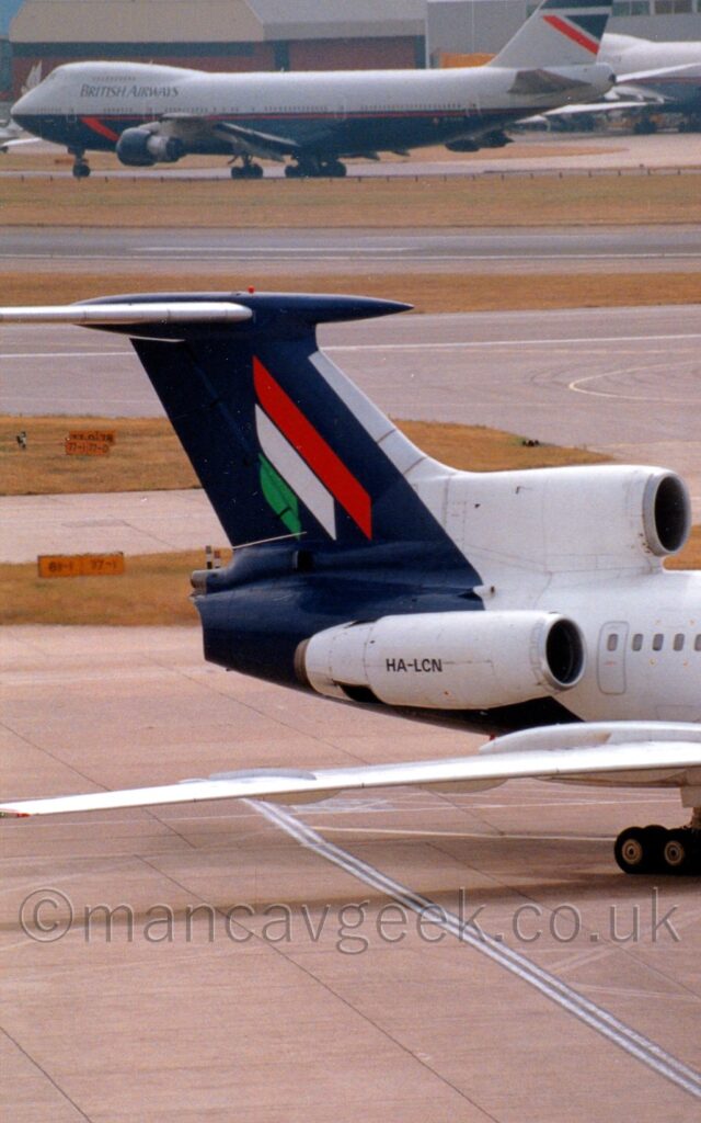 Close-up of the rear fuselage and tail of a 3 engined jet airliner, with the engines mounted on the rear fuselage, taxiing from left to right across a patch of grey apron. The plane is largely white, with a dark blue diagonal band wrapped around the rear fuselage and tail, with diagonal red, white, and green stripes, representing the Hungarian flag, and vaguely resembling an upper-case letter "M". The black registration "HA-LCN" is on the side of the white engine pods on the side of the rear fuselage. Grey apron fills the foreground, with grey taxiways and a runway separated by yellowing grass in the background, and a very large, 4 engined, grey airliner taxiing from right to left in the distance.