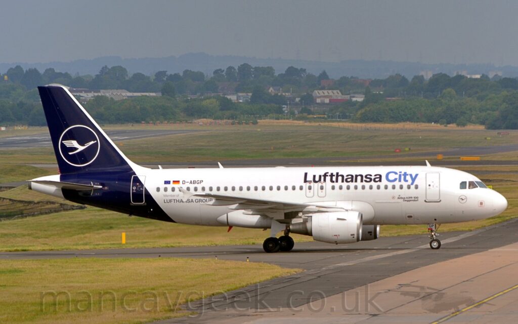Side view of a twin engined jet airliner taxiing from left to right. The plane is largely white, with dark blue "Lufthansa City" titles on the upper forward fuselage, the latter word in a lighter blue. The black registration "D-ABGP" is on the upper rear fuselage, next to small EU and German flags, with additional "Member of Lufthansa Group" text on the lower rear. The tail and rear fuselage are a dark blue, with the white outline of a circle containing a stylised white flying bird in the middle. A smaller, blue on white, version of this logo is under the cockpit windows on the plane's nose. Green grass and black and grey taxiways fill the foreground, as well as a large chunk of the background, leading up to trees and houses in the distance, under grey-blue skies.