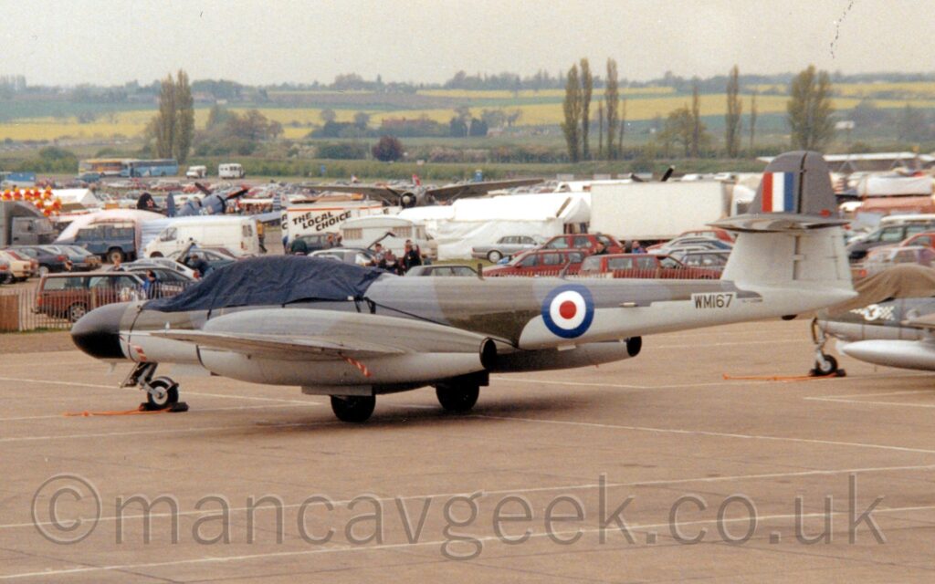Side view of a twin engined military fighter jet with the engines running through the mid-point of each wing, parked facing to the left and slightly away from the camera. The plane is mostly covered in a dark grey and brown camouflage scheme, with a lighter grey on the belly, bottom of the engine pods, and under the wings. There is a blue, white, and red roundel on the side of the rear fuselage, just ahead of the black serial "WM167" under the tail. There is a dark blue cloth cover over the tandem 2 seat cockpit. A large ground-level car park fills much of the background, with scattered white food trucks and tents, and the occasional vintage military aircraft on display. Trees and yellow and green fields in the distance meet grey sky.