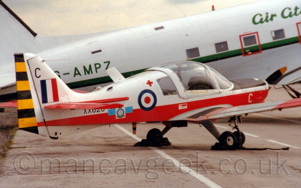 Side view of a white and red, single engined light aircraft parked facing to the right. The top half of the body is white, the lower half is red, and the horizontal tail-plane and ends of the wings are also red. There is a red, white, and blue roundel just aft of the over-sized, bulbous cockpit canopy, and a blue, whit, and red fin flash on the white tail. The serial "XX620" is on the rear fuselage in black, partially obscured by the tail-plane. A larger, white, World War 2-era airliner is parked in the background, with a green stripe running along the body, and the registration "G-AMPZ" in green on the rear fuselage.