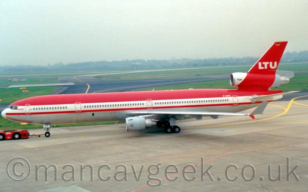 Side view of a 3 engined jet airliner being towed from right to left along a grey taxiway by a low red tug attached to the nosewheel by a long yellow pole. The plane has a top red half, with a thick white stripe running along the body, covering the passenger cabin windows, and a grey belly. There are small red "LTU" titles behind the cockpit windows, with a larger white version on the red tail, which also has a small German flag at the top. The inlet for the centre engine is white, the rest of the engine pod is red as it runs through the tail, with black text "TriJet" at the front. The white registration "D-AERZ" is on the upper rear fuselage, just forward of the rear door. Grey concrete apron fills the foreground, with black taxiways and a runway running through large grassed areas in the background. Trees and buildings in the distance are being swallowed up in misty grey sky.