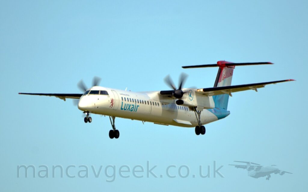 Side view of a white, high-winged, twin propellor-engined plane flying from right to left at a low altitude, with undercarriage fully extended from the rear of the engine nacelles, and flaps slightly extended from the rear of the wings, suggesting it is about to land. There is a red lion standing on it's rear legs on the lower forward fuselage, just forward of the door, with blue "LuxAir" titles below the passenger cabin windows just aft of the door. There is the blue outline of a circle containing a stylised blue flying bird on the sides of the engine nacelle. The tail is red and the top, blue at the bottom, and has a white stripe in between these colours, and is textured to look like cloth, so that it resembles a waving flag - specifically, the flag of Luxembourg. The same "flying bird in a circle" logo is at the top of the tail, except this time it is a white outline of a circle and a white bird. Pale blue sky fills the rest of the frame.
