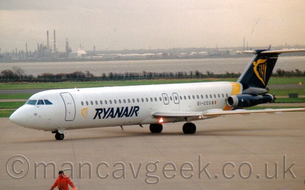 Side view of a twin engined jet airliner with the engines mounted on the sides of the rear fuselage, taxiing from right to left across a section of grey concrete apron. The plane is largely white, with dark blue "RyanAir" titles on the lower forward fuselage, slightly aft of a yellow, winged Irish harp. The black registration "EI-CCU" is on the lower rear fuselage, just ahead of a small Irish flag. A yellow band wraps around the rear fuselage, just ahead of the tail, while the tail, tail-cone, and engine pods on the rear fuselage are all dark blue, with a large yellow winged Irish harp in the middle of the tail. Grass fills the background, leading up to trees on the airfield perimeter, with a wide river beyond that, and an industrial landscape on the far bank, under grey skies.