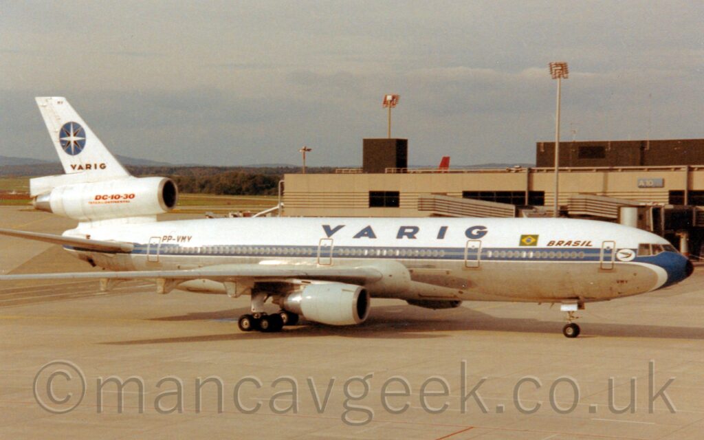 Side view of a white, 3 engined jet airliner taxiing from left to right. The plane has a blue stripe running along the body, covering the passenger cabin windows, and a bare metal belly. There are dark blue "Varig" titles, just aft of a small, green and yellow Brazilian flag and smaller blue "Brazil" titles. The black registration "PP-VMY" is on the upper rear fuselage, just in front of and below the tail. The white centre engine pods runs through the bottom part of the white tail, and has red text "DC-10-30" above smaller text "Intercontinental". There is black text "Varig" st the base of the top part of the tail, under a blue circle with a white and black 8 pointed compass in the middle, and the black letters "MY" right at the top. Grey concrete apron fills the foreground, with a sandy brown terminal building stretching across the background from the left. Trees in the distance meet layered cloudy sky with hints of blue at the top.