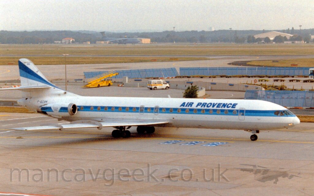 Side view of a sleek yet vintage-looking twin engined jet airliner, taxiing from left to right along a grey taxiway. The plane is largely white, with blue "Air Provence" titles on the upper forward fuselage, and a light and dark blue strip running along the body, covering the roughly triangular passenger cabin windows, as well as along the sides of the engine pods mounted on the sides of the rear fuselage. The tail is white, with the horizontal stabiliser mounted halfway up, and a light and dark blue stripe running along the top half, getting wider from a point as it moves rearwards. Grey concrete apron fills the foreground, with a couple of airport vehicles parked in the background, in the middle of an area partially surrounded by blue mesh metal fencing. Green grass beyond that has grey taxiways wending their way through, the grass leading up to a green, high-winged military jet transport and yellow buildings surrounded by trees in the distance, under hazy grey sky.