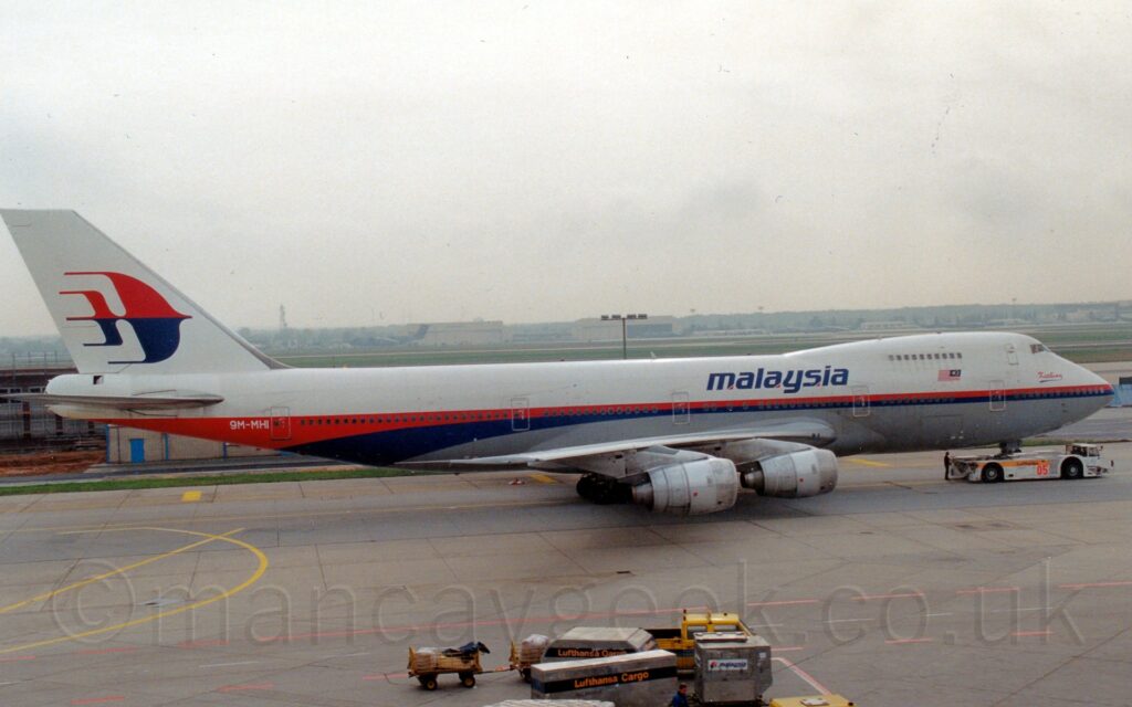 Side view of a very large, 4 engined jet airliner being towed from left to right by a low-slung white tug that is wrapped around the planes nose-wheel. The plane is mostly white, with a grey belly, and a red and blue stripe running along the body, getting wider towards the rear as it sweeps under the rear fuselage. Large blue "Malaysia" titles are on the upper forward fuselage, just aft of a small, red and blue Malaysian flag. The white registration "9M-MHI" is on the red stripe on the upper rear fuselage, while the tail is white, with a stylised, red and white image of an angel fish in the middle. Grey concrete apron marked with red and yellow lines fills the foreground, with grass in the background leading up to cream coloured hangars and green military transports in the distance, under cloudy white sky.