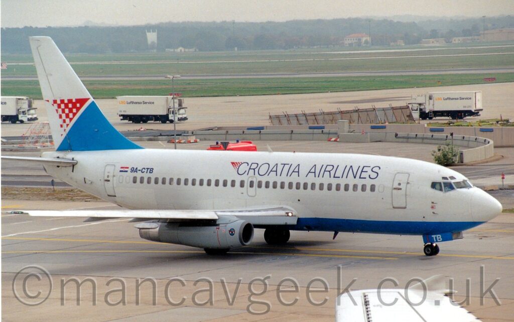 Side view of a twin engined jet airliner taxiing from left to right. The planer is mostly white with a blue belly, and black "Croatia Airlines" on the upper forward fuselage. The black registration 9A-CTB is on the upper rear fuselage, next to a small Croation flag. Grey concrete apron fills the foreground, with acres of green grass in the background, with trees vanishing in to haze in the distance.