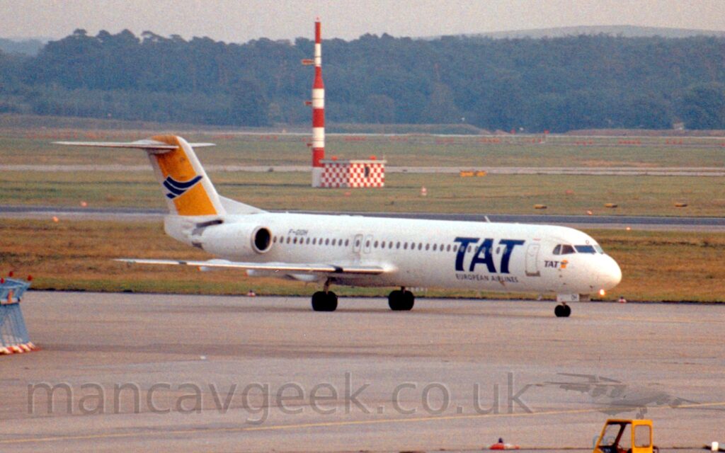 Side view of a twin engined jet airliner with the engines mounted on the sides of the rear fuselage, taxiing from left to right along a grey taxiway. The plane is mostly white, with large, blue, billboard-style "TAT" titles on the side of the forward fuselage, over smaller blue text "European Airlines". The tail is bright yellow outlined in white, with a stylised blue flying bird in the middle. A smaller version of this yellow and blue logo appears below the rearmost of the cockpit windows in the nose. The black registration "F-GIOH" is on the sides of the upper rear fuselage, just ahead of the white engine pods. Grey concrete apron fills the foreground, with grass in the background leading up to trees in the distance under grey sky.