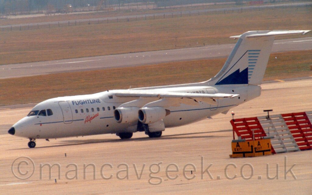 Side view of a high-winged, 4 engined jet airliner taxiing from right to left, with flaps extended from the rear of the wings, suggesting it is going out to a runway for takeoff. The plane is mostly white, with a thin blue stripe running along the body, overlaid with diagonal red "Alpine" titles below the passenger cabin windows just ahead of the wings, and blue "Flightlines" titles on the upper forward fuselage. The black registration "G-HWPB" is on the upper rear fuselage, partially obscured by the wing. The tail is white, with a blue and white stylised image of a snow-capped mountain, with short, light blue lines on it's leeward side. Grey concrete apron fills the foreground, with an angled red and white metal barrier on the right of the frame, while wide strips of green grass line a black runway in the background.