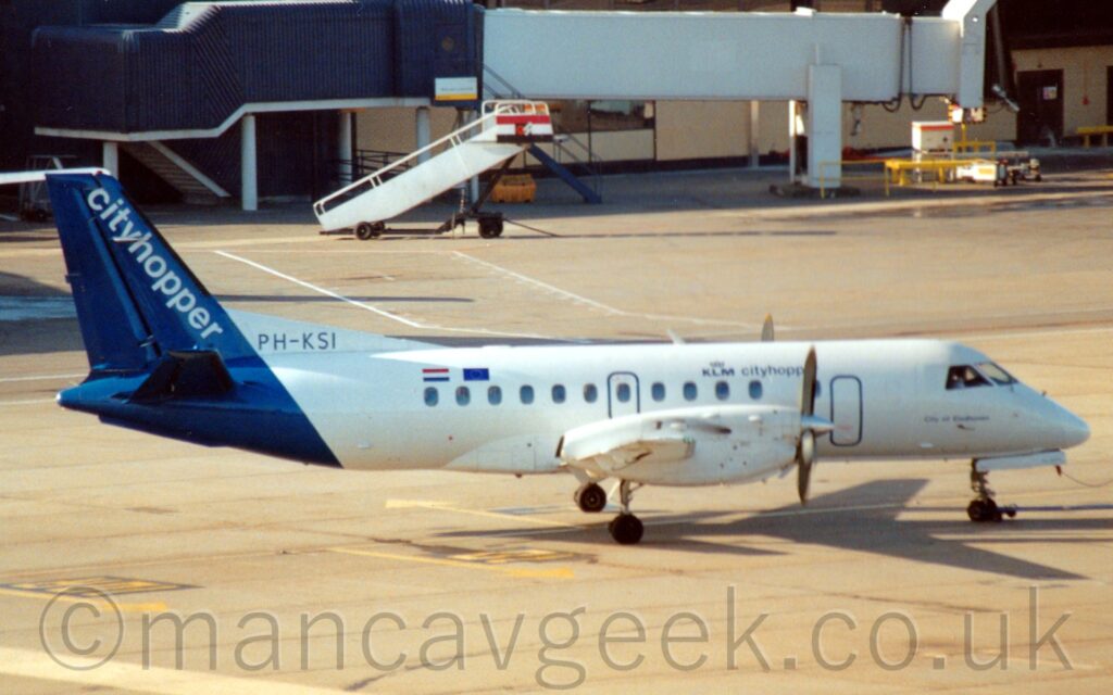 Side view of a twin propellor-engined airliner facing to the right but being pushed back to the left from it's stand by an out-of-frame tug attached to the nose-wheel by a long white pole. The plane is largely white, with "KLM CityHopper" titles on the upper forward fuselage, small text "City of Eindhoven" under the cockpit windows, and small Dutch and EU flags on the upper rear fuselage. The dark blue registration "PH-KSI" is on a white fillet on the upper rear fuselage at the front of the tail, while a dark blue band wraps around the tail and tailcone, with diagonal white "CityHopper" titles running along the leading edge. Grey concrete apron fills most of the rest of the frame, with a black and white airbridge hangs off the side of a sandy brown terminal building in the background.