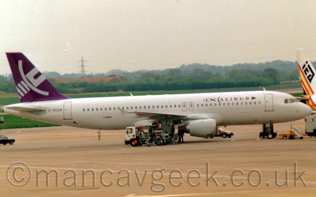 Side view of a white, twin engined jet airliner parked facing to the right on grey concrete apron. There are purple "Excalibur" titles on the upper forward fuselage, designed to vaguely resemble a long sword. There are small "A320" titles on the lower rear fuselage, and the purple registration "G-SCSR" on the upper rear, under the tail. The tail itself is purple, with the white hilt of a sword tilted up and to the right. Grey concrete apron fills the foreground, while large grassed areas lead up to trees in the distance, under grey sky. The white, black, red, and yellow tail of another jet airliner is peeking into frame from the right.