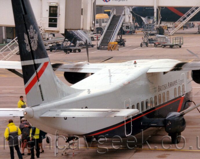Rear view of a squat and boxy, high-winged, twin propellor-engined airliner facing slightly to the right but mostly away from the camera. The plane has a grey upper half and dark blue lower, with a thin red pinstripe on the lower fuselage, ending in a half arrowhead at the front. There are black "British Airways Express" titles on the upper forward fuselage. The top half of the tail are dark blue, with a large grey heraldic crest in the middle. The lower half is grey, with a dark blue and red triangle at the rear. A group of people, some wearing bright yellow hi-viz jackets, are clustered around the left rear of the plane. Grey concrete apron fills most of the background, with A pair of white airbridges hanging off the side of a grey building on the left of the frame, and a larger jet airliner wearing the same livery nosing into frame from the right.