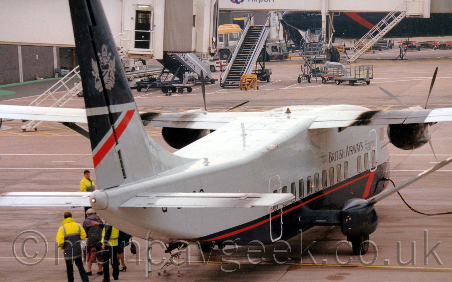 Rear view of a squat and boxy, high-winged, twin propellor-engined airliner facing slightly to the right but mostly away from the camera. The plane has a grey upper half and dark blue lower, with a thin red pinstripe on the lower fuselage, ending in a half arrowhead at the front. There are black "British Airways Express" titles on the upper forward fuselage. The top half of the tail are dark blue, with a large grey heraldic crest in the middle. The lower half is grey, with a dark blue and red triangle at the rear. A group of people, some wearing bright yellow hi-viz jackets, are clustered around the left rear of the plane. Grey concrete apron fills most of the background, with A pair of white airbridges hanging off the side of a grey building on the left of the frame, and a larger jet airliner wearing the same livery nosing into frame from the right.