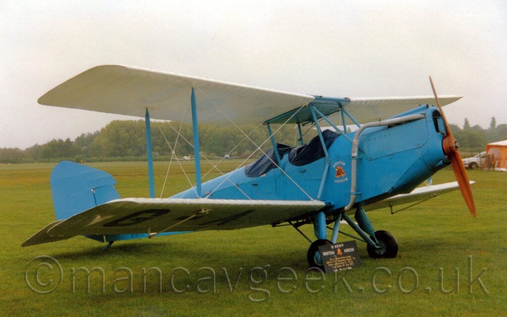 Side view of a pale blue biplane with white wings parked on grass facing to the right. The plane has 2 cockpits, covered with blue rubber covers to protect them from the elements. There is a red, yellow, and white triangle with white "Spartan Arrow" titles on top and below, under the front cockpit. Grass fills most of the frame, with trees in the distance, with a couple of other light aircraft parked in front, under grey skies.