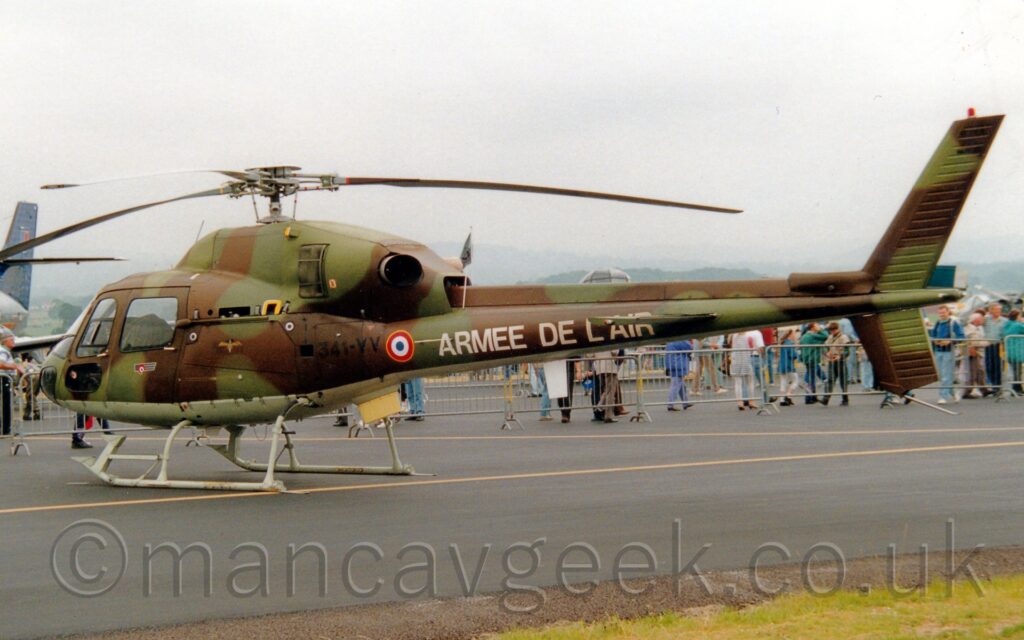 Side view of a small military helicopter parked facing to the left. The helicopter is covered in green and brown camouflage, with a pale green belly. There is black text "341-VV" on the side of the rear fuselage, next to a small blue, white, and red roundel outlined in yellow, and white "Armee De L'Air" titles on the sides of the tail boom. Black tarmac fills the foreground, with a patch of green grass in the bottom right corner. Several people can be seen walking past in the background, behind temporary metal barriers separating them from the planes on display. Rolling hills in the distance are being slowly swallowed up by misty grey sky.