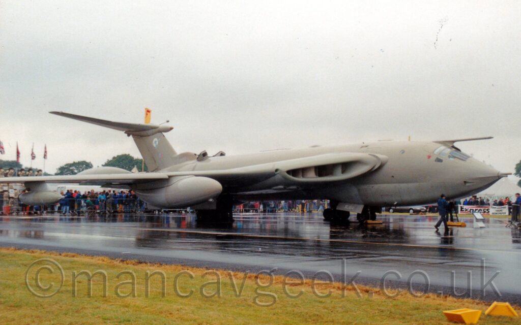 Side view of a surprisingly sleek, sandy brown, 4 engined jet bomber aircraft, parked facing to the right. The planes engines are buried deep in the wing root, with massive inlets feeding air into them. There are massive fuel pods under the wing, with a smaller pod on a pylon further out. There is a large probe mounted above the plane's cockpit. A man wearing a blue jacket and pants is walking in front of the plane's nose. The plane is parked on a sodden black taxiway, with grass lining the near side. In the background, crowds of people are gathered behind metal fences, wearing various types of rainwear. Grey skies fill the rest of the frame.