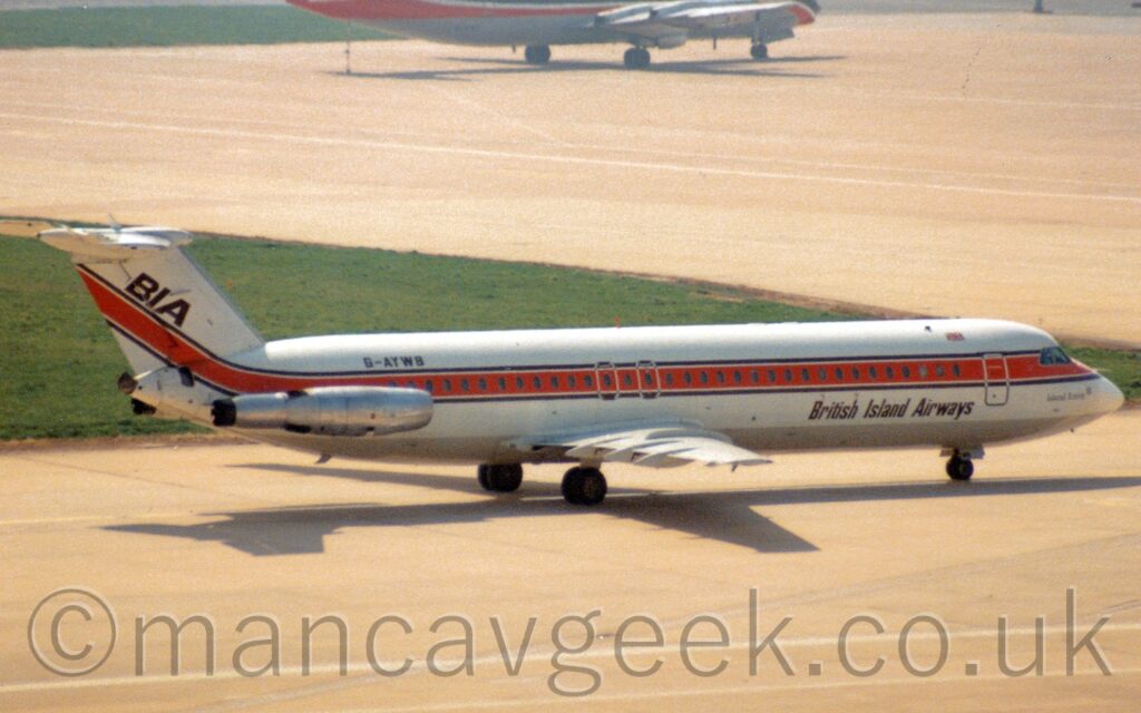 Side view of a twin engined jet airliner with the engines mounted on the sides of the rear fuselage, taxiing from left to right. The plane is mostly white, with a grey belly, and a thick orange stripe running along the body, outlined in black, covering the passenger cabin windows. There are black "British Island Airways" titles on the lower forward fuselage, and the black registration "G-AYWB" on the upper rear fuselage, above the bare metal engines. The orange stripe sweeps up into the tail, with diagonal black "BIA" text above it. Grey taxiway fills the foreground, with a large patch of grass on the left of the background, surrounded by more grey taxiway. The lower fuselage of a 4 propellor-engined, grey, red, and black airliner sits right at the top of the frame.