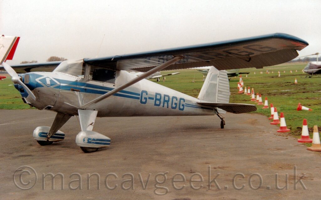 Side view of a high-winged single-engined light aircraft parked on a tarmac apron facing to the left. The plane is mostly in a bare metal finish, with a pair of blue stripes running along the body, getting narrower as they move to the rear, overlaid with the blue registration "G-BRGG" on the rear fuselage, which is repeated under the left wing. Another pair of blue stripes adorn the stripes on the spats over the main wheels. Struts support the wings, connected to the body just above the main gear legs. Grey tarmac apron fills the foreground, with a row of orange and white traffic cones separating that from muddy grass on the right of the frame. There are more light aircraft parked on grass in the background, under cloudy grey sky. Bright sunshine is casting a strong shadow of the plane on the ground.