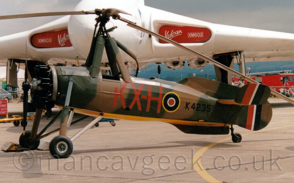 Side view of a 2 seat, single-engined gyrocopter parked in the static display park at an airshow, facing to the left. The plane is mostly covered in an olive green and brown camouflage scheme, with a bright yellow belly. The black serial "K4235" is on the rear fuselage, just aft of a blue, white, and red roundel outlined in yellow, itself just aft of the red letters "KX-H" under the rear cockpit. The 3 tails each have a large red, white, and dark blue fin flash. There are 4 supports around the open front cockpit, heading upwards, with a helicopter-style rotor at the top. A large white, delta-winged jet-engined military bomber aircraft dominates the background, with red covers with white text "Vulcan" and "XM603" in the engine inlets in the wing roots. Grey sky fills the rest of the frame.