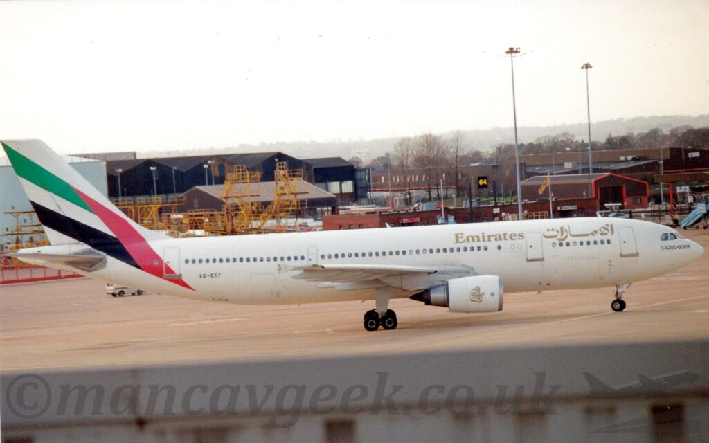 Side view of a twin engined jet airliner taxiing from left to right. The plane is mostly white, with gold "Emirates" titles on the upper forward fuselage, in an Arabic script at the front, and in English just forward of the wings. The back registration "A6-EKF" is on the lower rear fuselage. The tail and rear fuselage are white, with diagonal thick green, white, and black stripes connected to an almost vertically red bar vaguely resembling the flag of the United Arab Emirates if you squint. The top of a nearby grey wall stretches across the bottom of the frame, with grey concrete apron filling the rest of the foreground. Large duck egg blue fuel tanks are on the left of the background, with large black hangars, low brown buildings, and tall lighting poles filling the rest, and rolling hills in the distance, under grey skies.