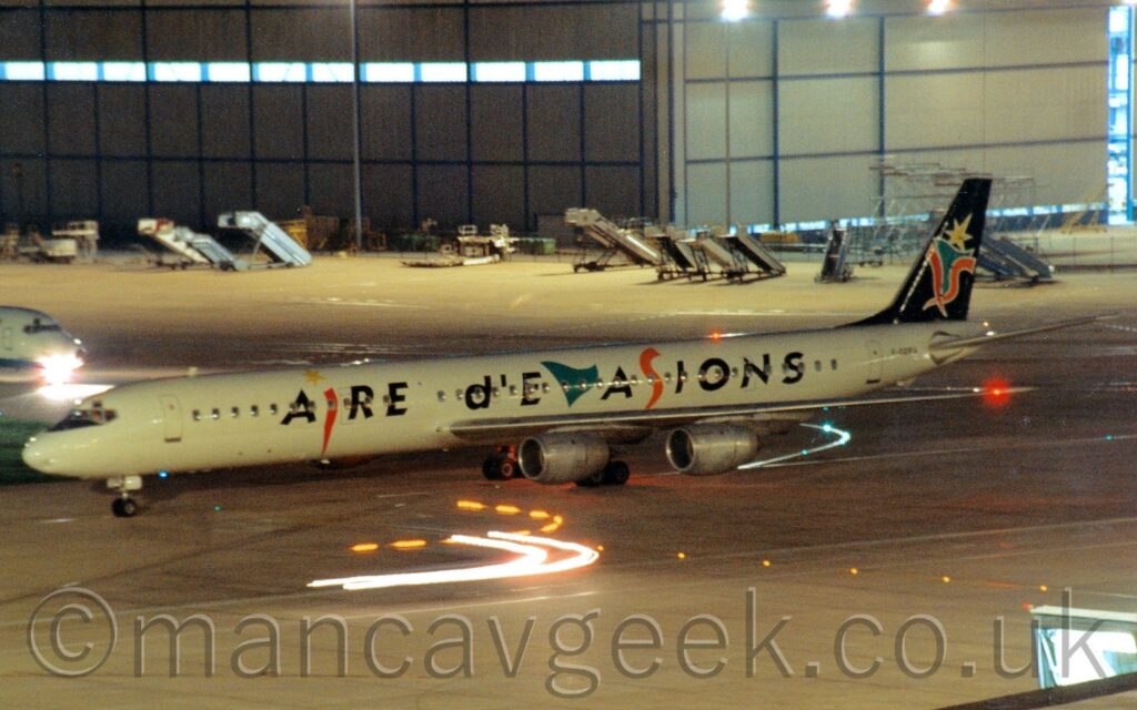 Long exposure night shot of a long, thin, white 4 engined jet airliner taxiing from right to left. There are black "Aire d'Evasions" titles along the body, with some letters replaced with red, green, and orange shapes, as well as a yellow star. These shapes are repeated and combined on the black tail. The black registration "F-GDRM" is on the side of the rear fuselage, under the tail. Red lights on the left wingtip and rear fuselage shine brightly in the dark. Grey concrete apron fills the foreground, with streaks of light from vehicles which have since moved out of frame, with white light shining inside an airbridge in the bottom right corner. More grey apron in the background leads up to a large grey hangar with bright white light shining inside, and mobile airstairs parked in front. The nose of another jet airliner is coming in to frame on the left of the background, with a bright white light shining from the nosewheel.