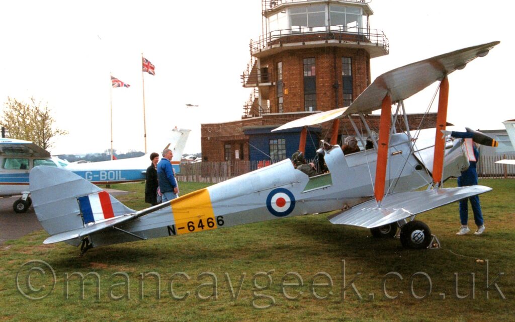 Side view of a single-engined, twin cockpit bi-plane trainer aircraft parked on grass facing to the right. The plane is in a mostly silver colour scheme, with a yellow band around the rear fuselage, overlaid with the black serial "N6466". There is a blue, white, and red roundel just aft of the wings, below the rear cockpit, and a blue white and red fin flash on the tail. Both cockpits are occupied, with people wearing heavy jackets and leather flying helmets. There is a flap open on the rear cockpit, to allow easier access for the occupant. There is a person standing at the front of the plane, partially hidden by the engine cowling, reaching up as if they are about to swing the propellor. A man and a woman are standing behind the rear of the plane, looking towards the person at the front. A white and blue light aircraft is sneaking out of frame on the left. A red brick control tower with a heavily glazed top floor fills the middle of the background, with a couple of tall flag poles towards the left of the frame, carrying Union Flags. Grey sky fills the rest of the frame.