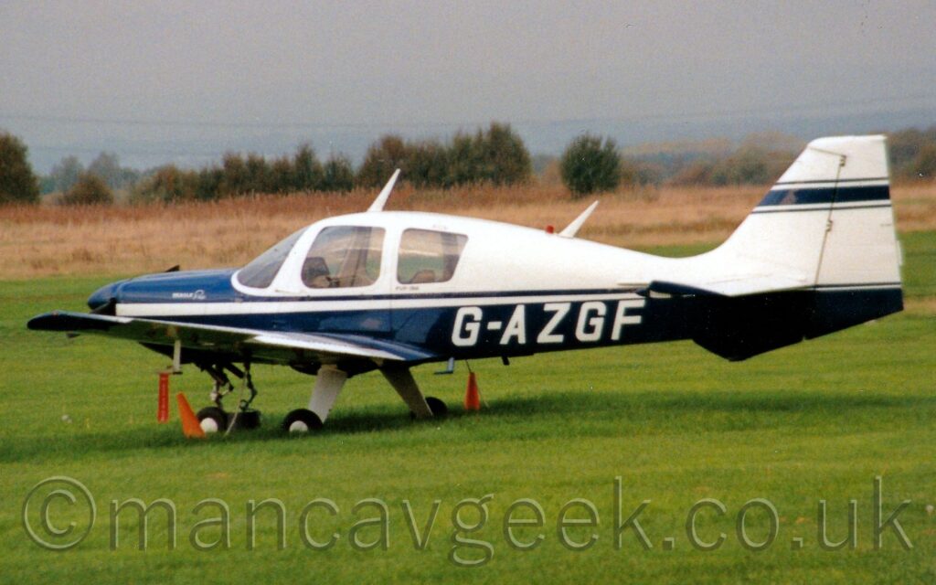 Side view of a single-engined light aircraft parked on grass facing to the left. The plane's lower half and nose is dark blue, with white upper surfaces and tail. The white registration "G-AZGF" is on the lower rear fuselage. Short green grass fills the foreground, with longer yellowing grass in the background leading up to trees in the distance under hazy grey skies