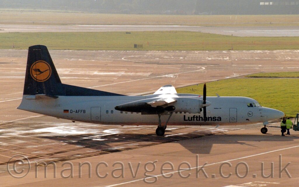 Side view of a high-winged, twin propellor-engined airliner facing to the right but being pushed back to the left by an out-of-frame tug attached to the nose-wheel by a long blue pole. The plane has a white body, with black "Lufthansa" titles on the lower forward fuselage, and the black registration "D-AFFB" on the side of the rear fuselage, next to a small German flag. The tail is dark blue, with a yellow circle in the middle containing highly stylised blue flying bird. Grey concrete apron fills the foreground, with a couple of people wearing hi-viz jackets by the plane's nose. More grey apron, a grey taxiway, and a black runway are in the background, separated by large grassed areas, with trees being swallowed up in haze at the top of the frame. Bright sunshine from the upper left is causing bright reflections on puddles in the background, and on top of the wings, as well as casting a strong shadow of the plane on the ground.